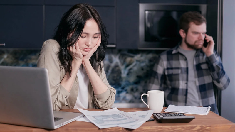 Two people in a kitchen. One is seated at a table with a laptop, papers, and a calculator; the other is standing in the background on the phone.